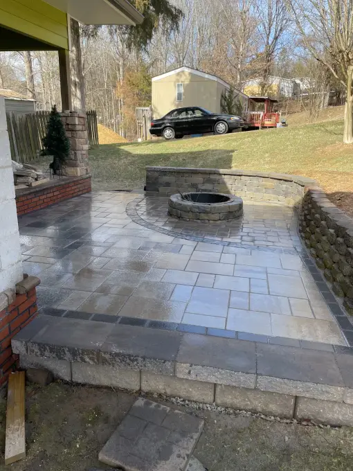 Paver patio with a round fire pit and low seating wall beside a yellow-sided home