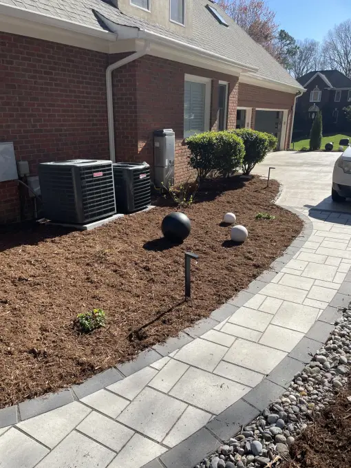 Paver walkway running beside a brick home past a fresh mulch bed with decorative stone spheres
